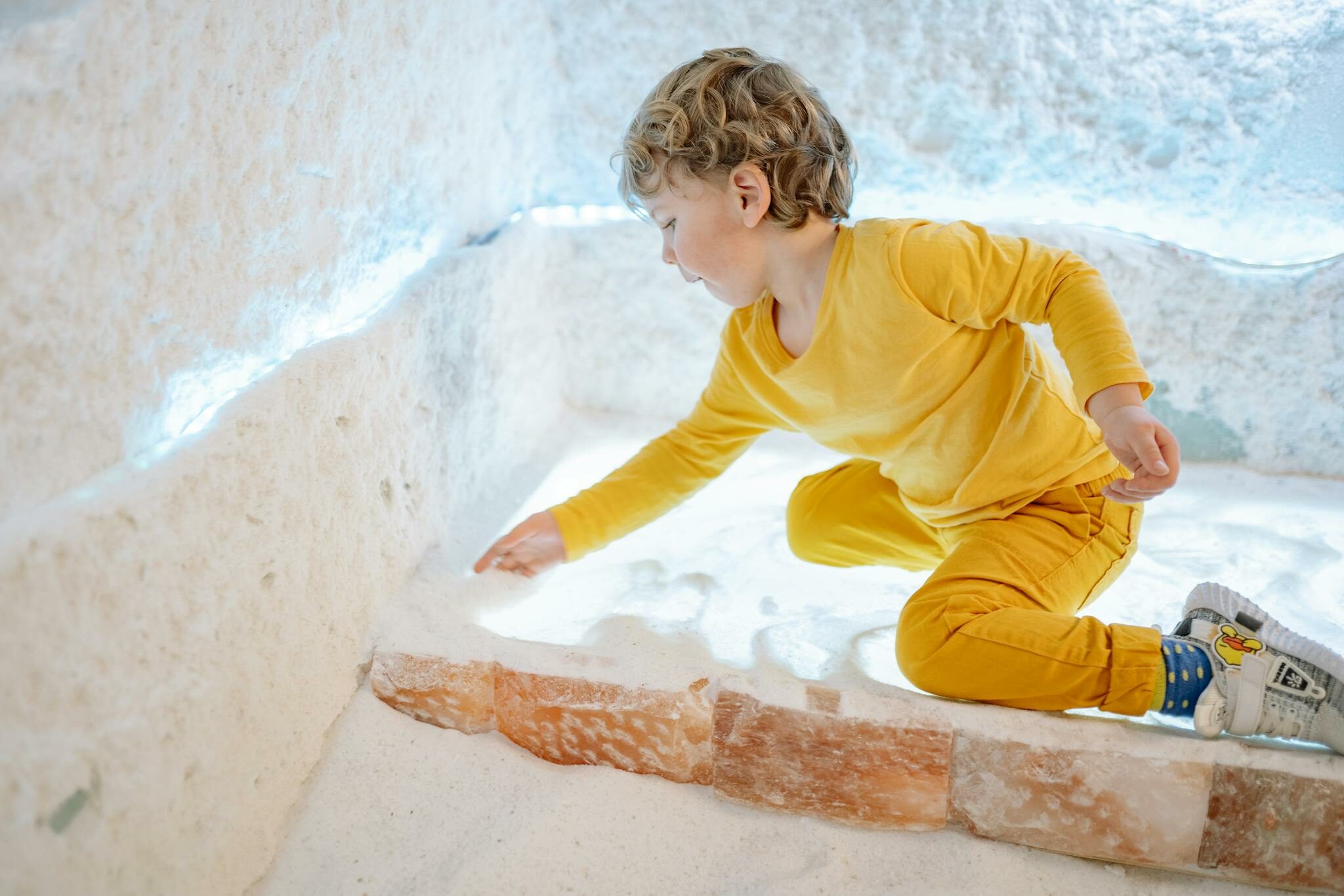 A young boy in a yellow outfit plays with salt in an illuminated indoor room.